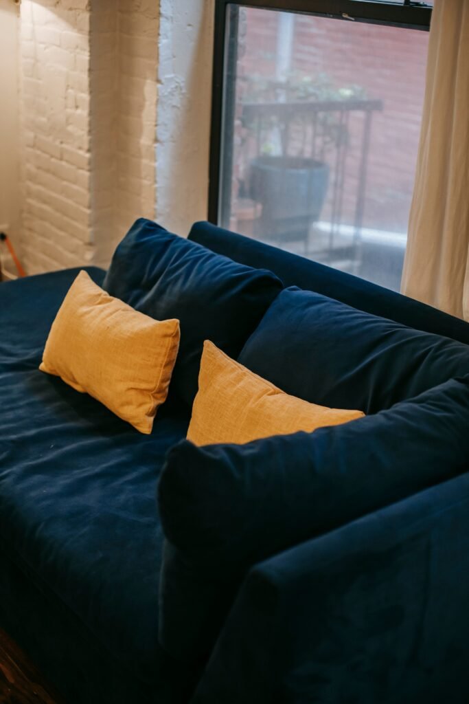 From above of blue soft couch with yellow cushions placed near window and white brick wall in light modern apartment